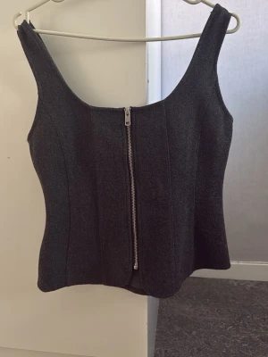 Mörkgrå korsettopp med dragkedja - Snygg mörkgrå korsettopp med breda axelband och dragkedja framtill. Toppen har en figurnära passform och är tillverkad i ett tjockare material som ger struktur. Perfekt att styla med jeans eller kjol för en trendig look.