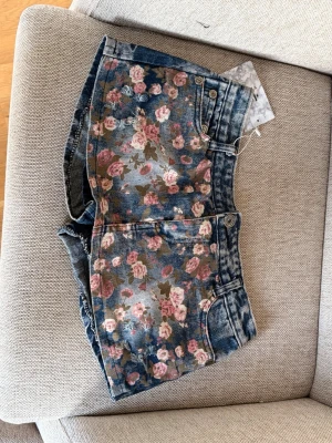 Vintage Blommiga jeansshorts med rosor - Snygga blå jeansshorts med rosa och grönt blommönster framtill och på ena bakfickan. Materialet är denim och shortsen har en cool, tvättad look. Köpte på en vintagebutik i Kina
