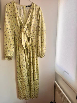 Gul blommig maxiklänning med knyt - Limegul maxiklänning med små blommor i lila och orange. Klänningen har långa ärmar med resår vid ärmsluten och en snygg knytning framtill. Tillverkad i ett mjukt, tunt tyg som ger ett flowy intryck. Perfekt för dig som gillar romantisk och feminin stil. Från Gina Tricot storlek 42