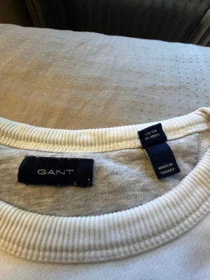 Två tröjor säljs tillsammans Vit sweatshirt från GANT - Tröjorna köps både två tillsammans och inte separata. Vit sweatshirt från GANT med broderad logga och texten 'American Sportswear' framtill. Tröjan har rund halsringning, ribbade muddar vid ärmslut och nederkant samt ett klassiskt collegeinspirerat tryck med sköld. Perfekt för en sportig och stilren look. 