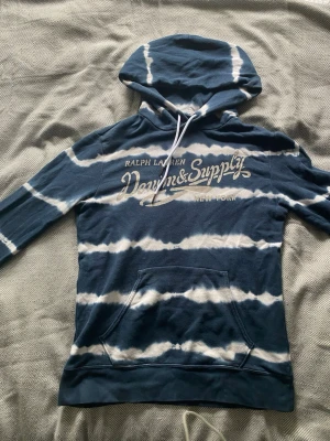 Blå tie-dye hoodie Denim & Supply - Snygg blå och vit tie-dye hoodie från Denim & Supply Ralph Lauren. Stor logga framtill, dragsko i huvan och mjukt material. Perfekt för en chill och avslappnad stil.