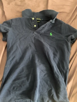 Marinblå piké från Ralph Lauren - Marinblå pikétröja från Ralph Lauren med klassisk krage, två knappar och grön broderad logga på bröstet. Modellen är 'The Skinny Polo' och har korta ärmar. Tillverkad i mjuk bomull för en skön känsla. Står storlek S men är nog närmre XS. Priset går att diskutera