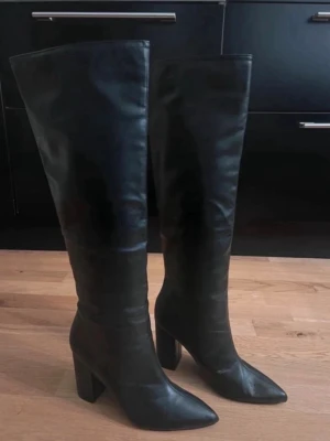 Svarta knähöga boots med klack - Snygga svarta knähöga boots med bred klack och spetsig tå. Tillverkade i ett glansigt material som liknar skinn. Perfekta för att lyfta vilken outfit som helst och ger en trendig siluett. Passar till kjol, klänning eller jeans.