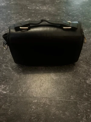 Svart boxig handväska i läderlook - Snygg svart handväska med boxig form och stilren design. Väskan har ett kort handtag upptill och detaljer i metall. Tillverkad i material med läderlook som ger en lyxig känsla. Perfekt för dig som gillar minimalistisk och modern stil. Inget är fel med väskan bara inte min stil 