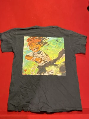 Svart T-shirt Thava Kavi med tryck - Cool svart T-shirt från trippy red med ett färgstarkt grafiskt tryck på ryggen som visar en tecknad figur med dreadlocks och tatueringar. På framsidan finns en liten orange detalj med siffran 14. Klassisk passform och rund hals.