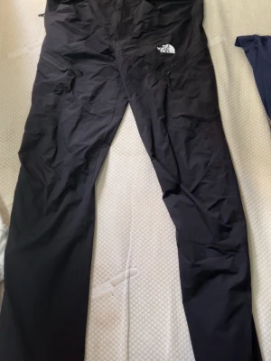 Svarta vindbyxor från The North Face - Säljer ett par svarta vindbyxor från The North Face med dragkedjor vid fickorna och logga på låret. Byxorna har en smal passform och är tillverkade i ett lätt, vindtätt material som passar perfekt för utomhusaktiviteter. Storlek 32/34