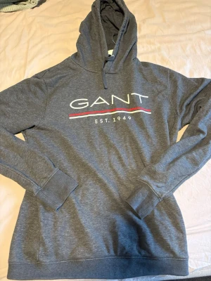 Grå hoodie från GANT med tryck - Snygg grå hoodie från GANT med stor logga och texten 'EST. 1949' på bröstet. Hoodien har huva med dragsko och ribbade muddar vid ärmar och nederkant. Tillverkad i mjukt bomullsmaterial och har en klassisk, avslappnad passform.