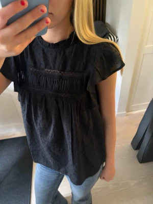Svart blus med spetsdetaljer - Säljer en svart blus med korta volangärmar och fina spetsdetaljer över bröstet. 