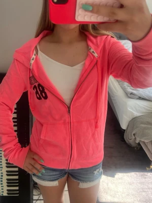 Rosa zip-hoodie från PINK - Säljer en snygg rosa hoodie från PINK med dragkedja och huva. På bröstet står det 'PINK 86' i svart tryck. Hoodien har fickor framtill och ribbade muddar vid ärmslut och nederkant. Perfekt för en chill och sportig look.