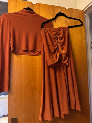 NA-KDxRebeccaStella set topp och kjol - Säljer ett snyggt rostbrunt set med en långärmad, croppad topp och en matchande kjol med rynkade detaljer baktill. Materialet är stretchigt och mjukt.