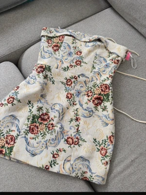 Blommig axelbandslös kortklänning - Unik axelbandslös kortklänning med vackert blommönster i rött, grönt och blått på beige botten. Klänningen har en figurnära passform och snörning i ryggen för justerbar passform. Perfekt för dig som vill sticka ut med en vintageinspirerad look.