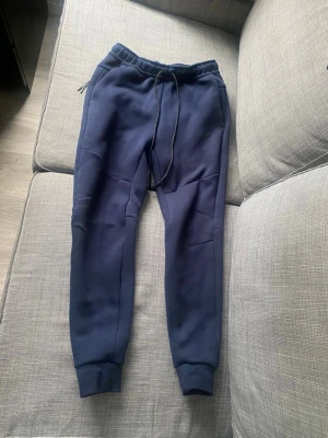Mörkblå Nike tech mjukisbyxor - Snygga mörkblå mjukisbyxor från Nike med dragsko i midjan och ribbade muddar vid bensluten. Byxorna har två sidofickor och en diskret Nike-logga på vänster ben. Perfekta för chill eller träning.