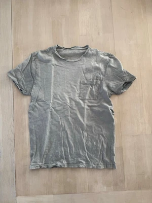 Grå basic t-shirt med bröstficka - En enkel grå t-shirt i bomull med korta ärmar och en liten bröstficka på vänster sida. Perfekt till vardags och lätt att matcha med andra plagg. Klassisk rund halsringning och avslappnad passform.