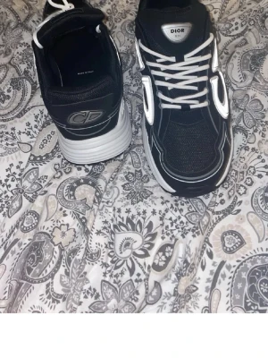 Svarta Dior B30 sneakers - Säljer ett par svarta Dior B30 sneakers med vita detaljer och stora CD-loggor på sidorna. Skorna har en chunky vit och svart sula, mesh och syntetmaterial i ovandelen samt vita skosnören. Perfekt för dig som gillar exklusiva och trendiga sneakers.