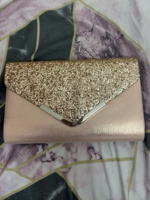Glittrig rosa kuvertväska  - Snygg kuvertväska i ljusrosa satin med glittrigt guldlock och silverdetalj. Väskan har en stilren och elegant design med triangelformad flik. Perfekt för att lyfta din outfit med lite extra glamour.