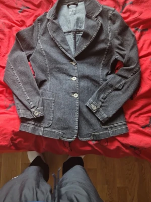 Svart jeansjacka från ril's - Snygg svart jeansjacka från ril's med klassisk kavajkrage, tre knappar framtill och två fickor. Jackan har markerade sömmar och knappdetaljer vid ärmsluten. Perfekt för dig som gillar en stilren och cool look.