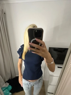 Mörkblå topp med volangärmar - Storlek 158/164-passar XS. Helt ny m prislapp! Trendig mörkblå topp med korta volangärmar och ribbad struktur. Sitter tight och har en snygg, figurnära passform. Perfekt att styla med jeans eller kjol för en cool look.