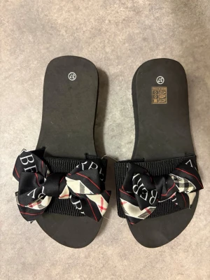 Svarta sandaler med text, mönster och rosett på - Svarta sandaler med text, mönster och en rosett på. Supermjuka och härliga att ha på sig. Aldrig använda, man ser plasttråden mellan tofflorna är kvar.