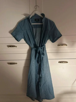 Blå jeansklänning från Object - Snygg blå långklänning i jeansmaterial från Object. Klänningen har korta ärmar, knappar framtill och ett matchande knytband i midjan för en schysst siluett. Perfekt till sneakers eller boots för en cool look.