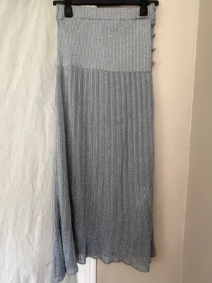 Lång silverfärgad ribbad kjol, vintage - Säljer en lång, ribbad kjol i glittrigt silver med knappar längs sidan. Kjolen har en hög midja och ett mjukt, stretchigt material som ger en snygg passform. Perfekt för dig som vill sticka ut med en unik och trendig look.