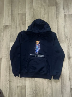 Svart hoodie Polo Bear Ralph Lauren - Svart hoodie från Ralph Lauren med den ikoniska Polo Bear-trycket på bröstet. Björnen har solglasögon, randig tröja och jeansjacka. Hoodien har huva med dragsko och en stor magficka framtill. Perfekt för dig som gillar streetwear och klassiska märken.
