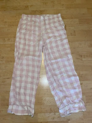 Rutiga pyjamasbyxor i rosa och vitt - Mysiga pyjamasbyxor med rosa och vitt rutmönster. Byxorna har resår i midjan och raka ben med en avslappnad passform. Perfekta för chillkvällar hemma. Materialet känns mjukt och luftigt, troligen bomull eller bomullsblandning.