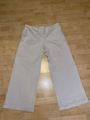 Vida beige kostymbyxor från H&M - Snygga beige kostymbyxor från H&M med vid passform och hög midja. Byxorna har klassisk dragkedja och knapp framtill. Tillverkade i ett mjukt, vävt material som ger en stilren look. Perfekta för dig som gillar en avslappnad men dressad stil.