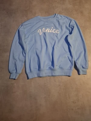 Ljusblå sweatshirt med Venice-tryck - Ljusblå sweatshirt med rund hals och ribbade muddar. Framtill står det 'Venice' i vit text. Tröjan har en relaxed fit och är gjord i mjukt bomullsmaterial. Perfekt för en chill och enkel stil.