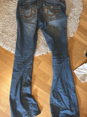 Blå bootcut jeans med broderade fickor - Säljer ett par blå bootcut jeans från True Religion med snygga slitningar och broderade detaljer på bakfickorna. Jeansen har klassisk femficksmodell och är tillverkade i denim med lätt stretch. Perfekta till skolan/jobbet men också fest! 