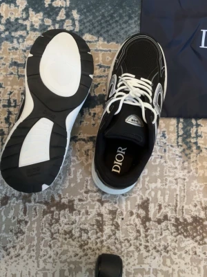 Svarta Dior sneakers med vit sula - Snygga svarta Dior sneakers med vit och svart tjock sula, vita detaljer och vita skosnören. Ovandelen är i mesh och läder med tydliga Dior-loggor på plösen och innersulan. Perfekta för dig som vill ha en exklusiv och trendig look.