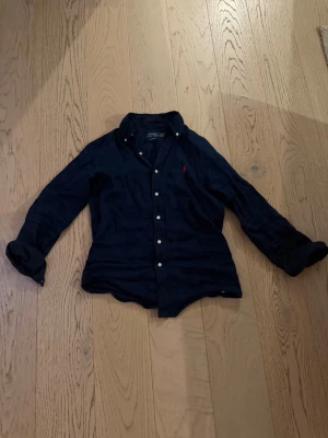 Mörkblå linneskjorta Polo Ralph Lauren - Klassisk mörkblå skjorta från Polo Ralph Lauren i linne med custom fit. Skjortan har långa ärmar, knappar framtill och den ikoniska röda loggan broderad på bröstet. Perfekt för en stilren och avslappnad look.