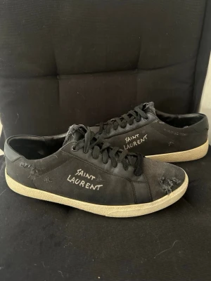 Saint Laurent skor - Riktigt snygga svarta sneakers från Saint Laurent med vit logga broderad på sidan. Skorna har slitna detaljer, svarta skosnören och vit sula. Materialet är en mix av mocka och läder vilket ger en cool kontrast. Perfekta för dig som gillar en edgy streetstyle. Skriv vid frågor!