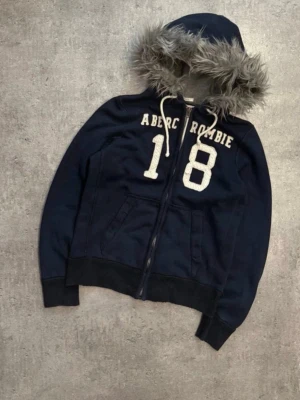 Marinblå hoodie Abercrombie & Fitch med päls  - Snygg marinblå hoodie från Abercrombie & Fitch med dragkedja, stora vita siffror '18' på bröstet och texten 'Abercrombie'. Huva med grå fuskpäls och vita dragsnören. Mjuk bomullsblandning och två fickor framtill. Perfekt för en chill och avslappnad stil. Fet og hoodie med päls 