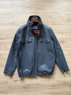 Loro Piana Jacka - Stilren jacka från Loro Piana med hög krage och dragkedja framtill. Två bröstfickor med lock och två sidofickor. Insidan är fodrad i rostbrunt tyg och har en innerficka. Perfekt för dig som vill ha en klassisk men modern look.