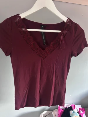 Vinröd topp med spets  - Vinröd topp med v-ringning och dekorativ spets runt halsen. Kortärmad modell i mjukt material som sitter snyggt på kroppen. Perfekt att styla med jeans eller kjol för en trendig look.