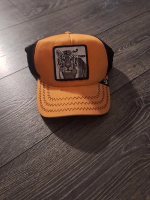 Orange keps från The Farm Tiger - Snygg orange keps från The Farm med svart mesh baktill och ett broderat tiger-märke framtill. Kepsen har justerbar snapback och coola zigzag-sömmar på skärmen. Perfekt för dig som vill sticka ut med djurmotiv och streetwear-vibe.