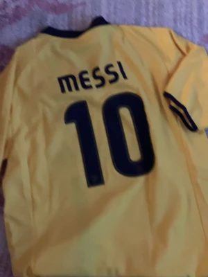 Gul FC Barcelona Messi tröja - Snygg gul FC Barcelona matchtröja från Nike med Messi och nummer 10 på ryggen. Tröjan har klubbmärke, Unicef-logga och en vertikal röd-blå rand på framsidan. Perfekt för dig som älskar fotboll och vill representera Barca.