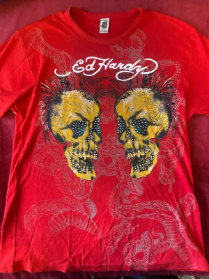 Vintage Ed Hardy t-shirt - Cool röd t-shirt från Ed Hardy med stort tryck av två gula dödskallar med punkfrisyr och nitar på framsidan. T-shirten har rund hals och korta ärmar. Materialet är mjuk bomull och trycket har detaljer i svart och vitt.