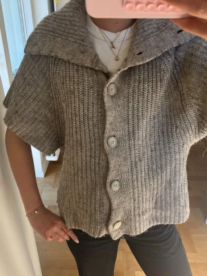 Beige stickad kofta med stora knappar - Kortärmad stickad kofta i beige med stora vita knappar framtill och bred krage. Grovstickad struktur som ger en avslappnad look. Perfekt att slänga över en t-shirt för extra lager. Märke syns ej tydligt på bilden.