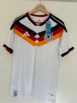 Tysklands matchtröja Adidas M - Säljer en vit Tyskland matchtröja från Adidas med klassiskt mönster i svart, rött och gult över bröstet. Tröjan har korta ärmar, svart och röd krage samt officiellt landslagsmärke på bröstet. Tillverkad i lätt och ventilerande climalite-material.