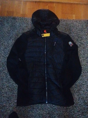 Para jumpers Svart vindjacka - Snygg svart vindjacka från Parajumpers med huva och dragkedja framtill. Jackan har vadderade paneler, en bröstficka med dragkedja och den klassiska Parajumpers-loggan på ärmen. Orange detalj vid halsen och kontrastsömmar på axlarna. Perfekt för blåsiga dagar.