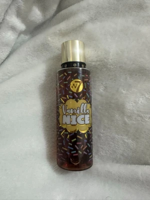 W7 Vanilla Nice Body Mist - En söt body mist från W7 med doft av vanilj. Flaskan är genomskinlig med färgglada strössel på etiketten och har ett guldigt lock. Perfekt för dig som gillar gourmanddofter och vill ha en lekfull parfym i badrumshyllan.