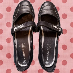 Svarta ballerinaskor från Head - Snygga svarta ballerinaskor från Head med vit kontrastsöm och kardborreband över foten. Skorna har rundad tå, dekorativa nitar på sidan och logga på bandet. Perfekta för en sportig och avslappnad stil.