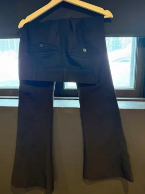 Svarta bootcut kostymbyxor - Snygga svarta kostymbyxor med utsvängda ben och två bakfickor med lock och knapp. Klassisk passform med hällor för skärp och knappstängning fram. Perfekta för en stilren look. 
