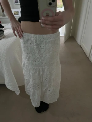 Vit långkjol med broderade blommor - Säljer en vit långkjol med elastisk midja och broderade blommor i tyget. Kjolen har en luftig och flowy passform, perfekt för varma dagar. Materialet känns som bomull och ger en romantisk vibe. Supersnygg att styla med crop tops eller linnen. Säljer på grund av att den är för kort 