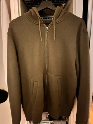 Olivgrön zip hoodie med huva - Säljer en stilren olivgrön hoodie med dragkedja framtill och två fickor. Hoodien har en klassisk huva med dragsko och ribbade muddar vid ärmslut och nederkant. Perfekt för en avslappnad look och enkel att matcha med jeans eller joggers. Storleken är L men passar för M också