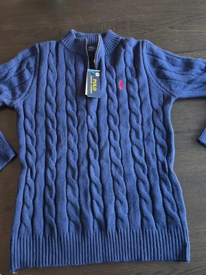 Blå kabelstickad tröja Polo Ralph Lauren - Kabelstickad blå tröja från Polo Ralph Lauren med halv dragkedja och röd broderad logga på bröstet. Tröjan har ribbade muddar och hög krage, perfekt för dig som gillar klassisk och stilren design.