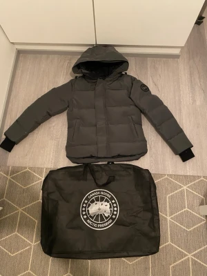 Mörkgrå dunjacka från Canada Goose - Snygg mörkgrå dunjacka från Canada Goose med huva och diskret logotyp på ärmen. Jackan har två stora framfickor, dragkedja med vindslå och ribbade muddar vid ärmsluten. Perfekt för kalla vinterdagar och riktigt varm.