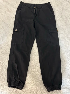 Svarta cargobyxor med resår - Svarta cargobyxor med stora fickor på sidorna och resår vid bensluten. Byxorna har knappstängning och snörning i midjan samt bakfickor med lock. 