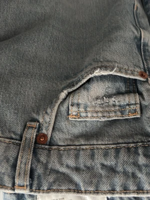 Blåa slitna jeans med femficksdesign - Ett par klassiska blå jeans med femficksdesign och slitna detaljer runt fickorna. Jeansen har en vintage look med ljus tvätt och synliga sömmar. Perfekt för dig som gillar en avslappnad och trendig stil.